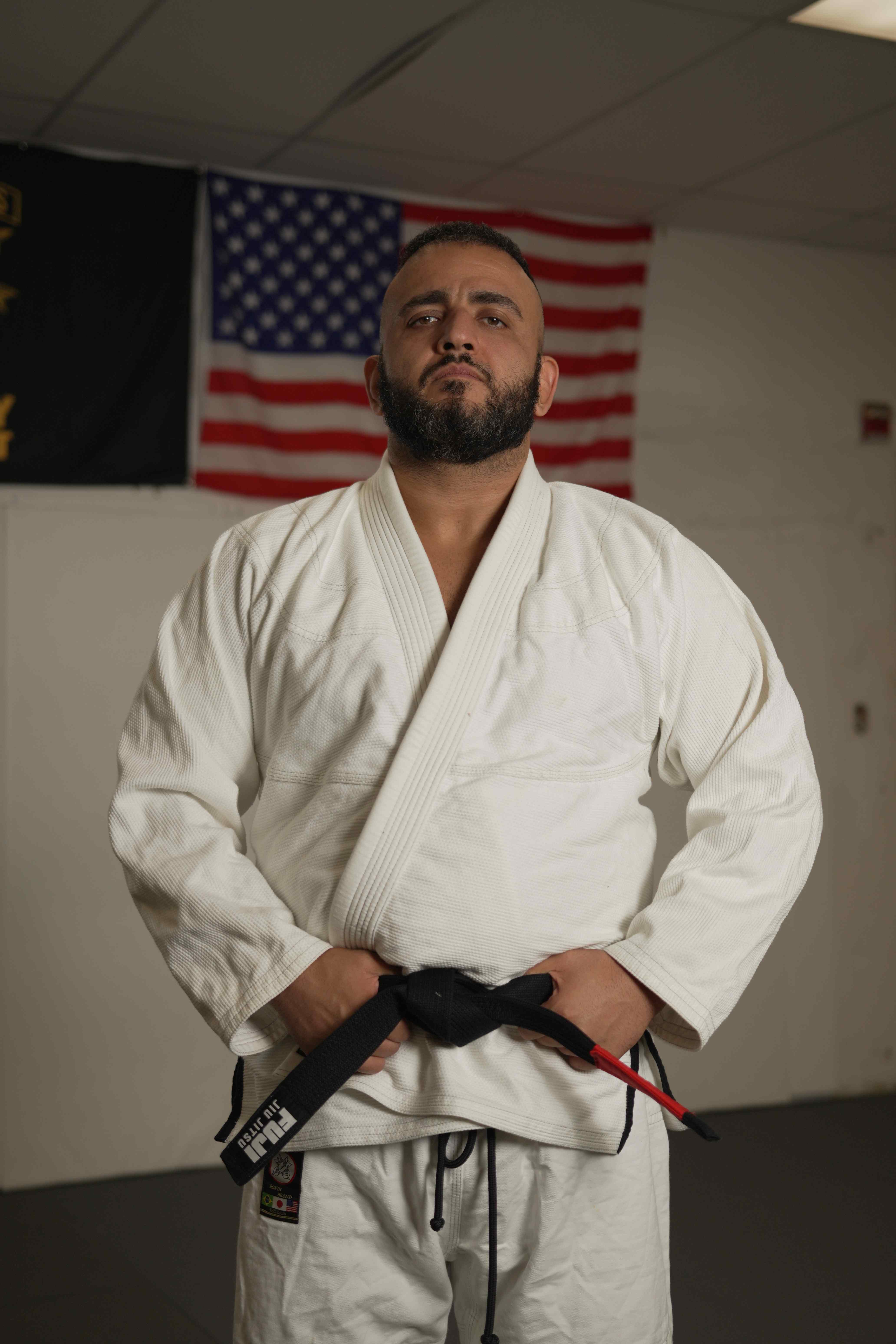 Mike Rakshan, founder and head coach of Immortals Jiu Jitsu, in his gi and black belt in front of the American flag