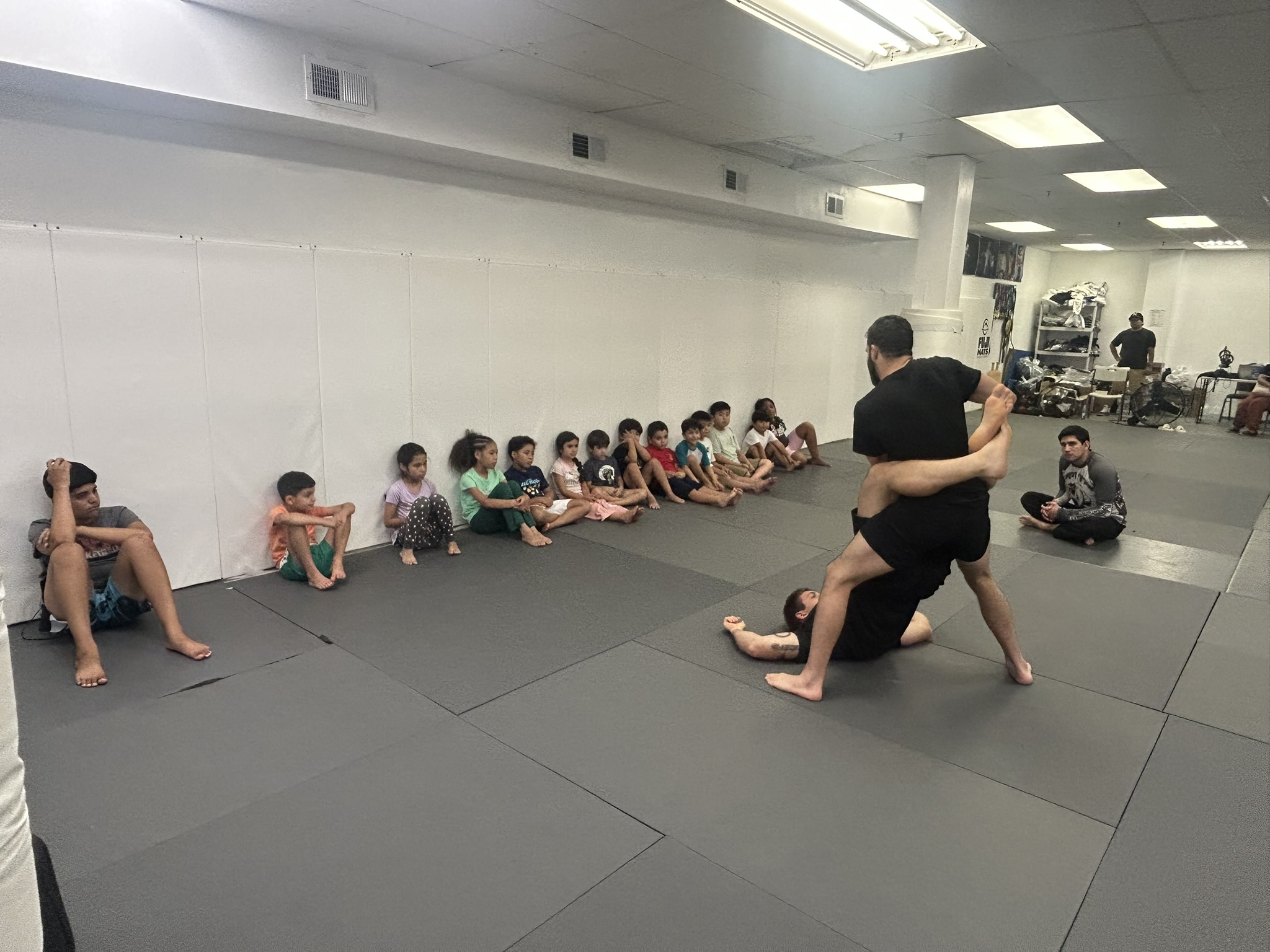 Young students sitting along the wall watching an Immortals coach demonstrate a jiu jitsu technique during a kids class.