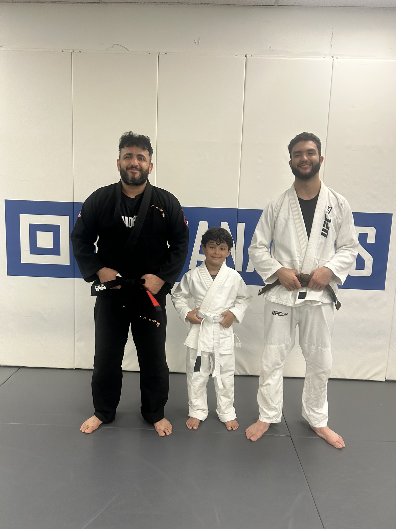 A young student in a white gi standing between two Immortals coaches after a kids jiu jitsu class.