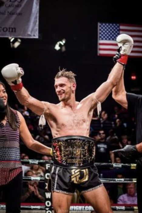 Ognjen Topic celebrating a Muay Thai title win in the ring with the championship belt and gloves raised.