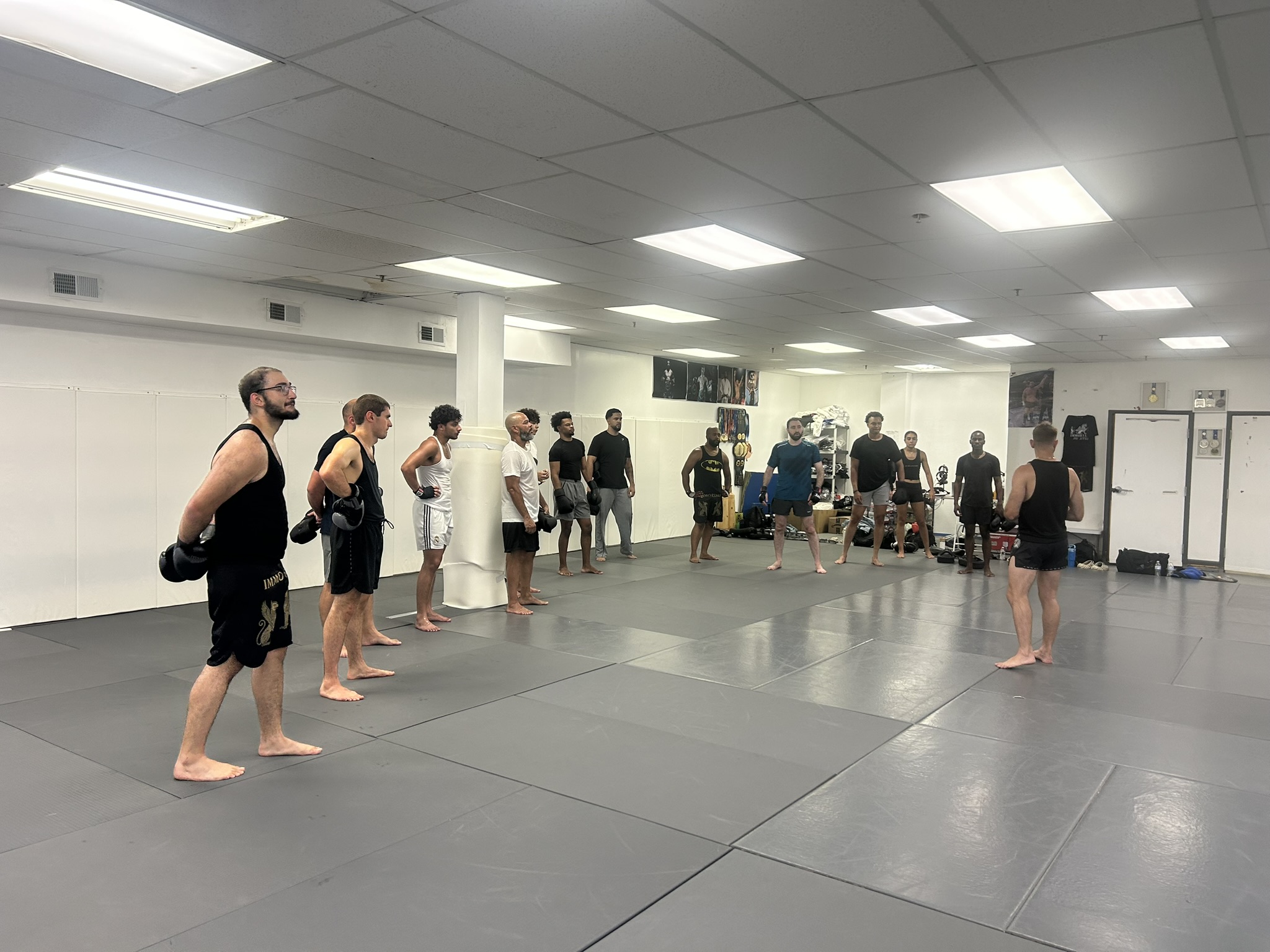 An adult Muay Thai class lined up on the mats with boxing gloves as the coach leads the session.